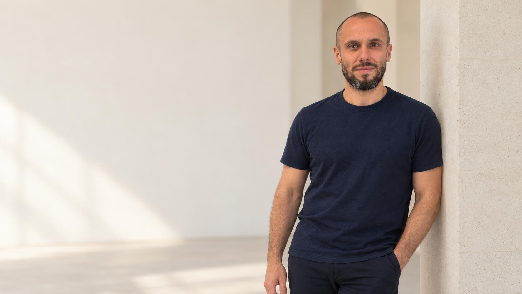 Andreas Köhn, standing against a stone column in a bright interior, wearing a navy T-shirt.